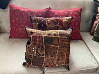 Four throw pillows on a couch, showing two red pillows with mirror detailing and two earthy-toned patchwork pillows with mirror accents and tassels.