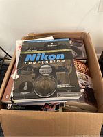 Box of books showing 'The New Nikon Compendium' with camera images on cover and several other books underneath.