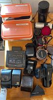Overview showing two orange Sunpak flash cases, Sony VCR, Nikon and Canon flashes, Porroflex viewfinder, Miranda lens, Topcon lenses, leather pouches