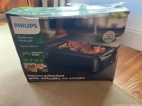 Photo showing front side of the box with Philips branding, image of the indoor grill and grilled food items, smoke-less infrared technology indicated on packaging.