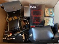 Box containing various camera accessories including Vivitar 283 flash, Tasco slide film, Canon case, and other items