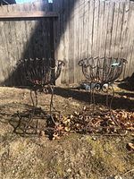 Two metal outdoor planters standing on dirt with a wood fence background showing the full view of each with their open wire basket design and legs.