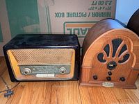 Two vintage radios on wooden floor, Rekord Junior 196 on left with black case, Philco wooden cathedral style radio on right.