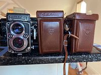 Three cameras side by side on counter: uncased Rolleiflex at left, two leather-cased cameras at center and right