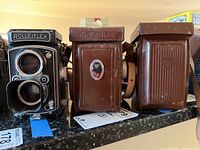 Three twin-lens reflex cameras side by side on countertop: left camera without case with metal lens hood, middle and right cameras in brown leather cases