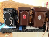Three Rolleiflex cameras side by side (one loose, two in brown leather cases) on shelf