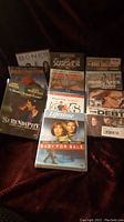 Photo showing an assortment of sealed DVDs on a dark surface, titles include Serendipity, Paddington, Bones, The Debt and others.