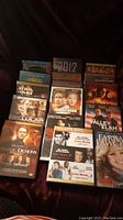 Photo showing collection of 13 DVD movies arranged in three rows on a dark fabric surface, with visible titles and cover art.