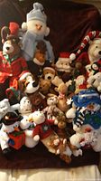 Photo showing a large assortment of Christmas-themed stuffed animals including various snowmen, reindeer, Santa Claus, and elves arranged on a dark surface.