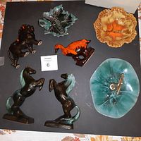 Top-down image showing all seven Blue Mountain Pottery pieces on black background: two horses, orange goose, teal Canada maple leaf plate, orange dish, teal dish with central decoration.