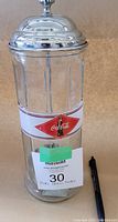 Front view of clear glass Coca-Cola straw dispenser with red and white logo and metal lid with knob, next to a pen for scale.
