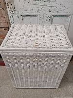 Front view of a square white wicker clothes basket with a lid and latch closure placed against a distressed white door.