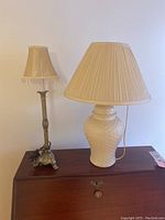 Photo showing two desk lamps on a wooden surface: one with a cast metal ornate base and fringe trimmed fabric shade, the other with a white ceramic basket weave base and pleated shade.
