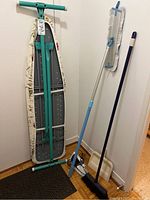 Full view of the ironing board fully folded and standing, the wet dust mop with blue and white handle, broom with blue handle, and beige dustpan all placed in a corner.