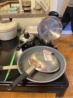 Two Paderno frying pans stacked with black handles visible, alongside stainless steel sauce pans and a white container in the background.