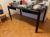 Full view of the black painted wood table showing the rectangular top with integrated drop leaf on the near side, four tapered legs, set in a room with parquet flooring and some items on table