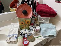 Overview of bathroom accessories lot on white countertop, showing towels, toothbrush holder, small jars, hand mirror, and vintage bottle in sunflower container.