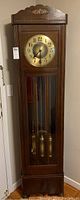 Full view of tall wooden grandfather clock against a wall showing the overall design, glass door and visible pendulum and weights.