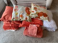 Top view of the pile including floral tulip cushions, solid orange cushions, orange towels, and throws
