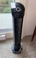 Full view of the Seville Classics tall tower fan with black and silver body on carpeted floor near window.