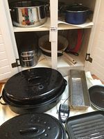 Photo depicting various cookware items including two large black speckled roasters with lids, a loaf pan, and a digital cooking thermometer, arranged on a countertop and inside a cabinet.