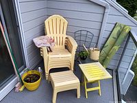 Stacked beige Adirondack-style plastic chairs with one chair showing a floral patterned cloth draped over the arm. Yellow planter and broom nearby. Yellow folding table, black metal stand and outdoor mat are visible.