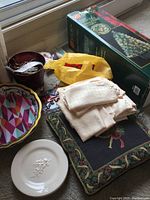 Assorted seasonal decor on windowsill including boxed fiber optic Christmas tree, folded cloth linens, a holiday pillow, ceramic plates, and a candle.