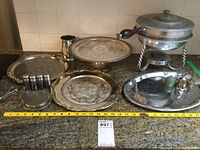 Photo showing various silver electroplated and metal trays, some tarnished, displayed on a countertop along with a metal chafing dish with wooden handle and small silver accessories.