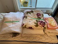 Full view of folded twin bed linens set on mattress near window, showing mattress cover, floral duvet, beige blanket, and folded linen with labels.