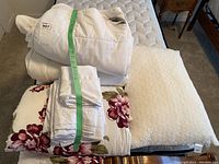 Photo showing folded white mattress cover on top, white duvet underneath, floral blanket with burgundy flowers, folded white bedding, and textured cream pillow on a twin mattress.