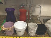 Lot showing all vases and planters of mixed materials and colors arranged together on countertop with measuring tape for size reference.