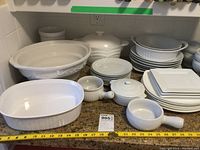 White ceramic kitchenware arranged on countertop including casserole dishes, plates, bowls, ramekins, and spoons. Shows the lot contents clearly.