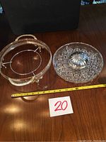 Photo showing the glass serving dish with silver lid next to the round metal holder with handles, both resting on a wooden surface with measuring tape and a lot number card.