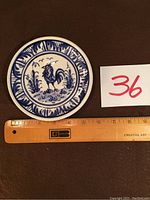 Top view showing full plate design with blue rooster and decorative border, next to a ruler for size reference.