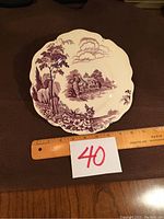 Full view of the Avon Cottage collector plate showing the purple transferware design and scalloped edges, with a ruler for scale and a lot number card.