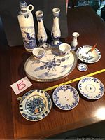 Full set of 13 Delft ceramic pieces displayed on wooden table showing various shapes and hand painted blue floral and windmill motifs. Includes tall bottles, vases, plates, tray, ashtray, small plates and cups.