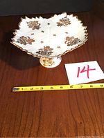 Full view of white raised candy dish with scalloped edges and gold rose design on wooden table with measuring tape.
