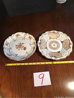 Photo showing two antique German made china plates with gold accents on a wooden surface with measuring tape for scale.