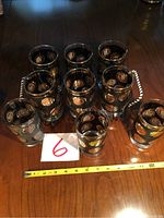 Group view of nine clear glasses decorated with gold coin embossed USA symbols arranged on a wooden table with a measuring tape showing size and a numbered label 6.