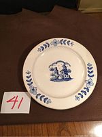 Full view of round ceramic collector plate with blue floral border and central image of two houses with archway.