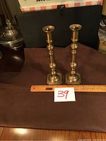 Photo showing the pair of tall brass candle holders on a dark surface with a ruler and lot number 39 visible for scale.