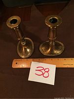 Pair of brass candle holders displayed next to a ruler showing their height about 6 inches. Candle holders have round bases and cylindrical tops with noticeable patina.