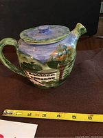 Side view of handmade ceramic teapot with painted houses and greenery, showing lid and spout, approximately 6 inch size