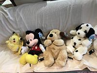 Four plush character and animal backpacks on a white background: Pikachu, Mickey Mouse, golden retriever dog, and a black and white spotted dog.