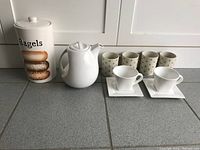 Photo showing the ceramic bagels container, white coffee pot, two white square coffee cups with matching saucers, and three beige small cups with twig pattern.