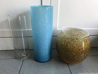 Three hand made glass vases side by side on tiled floor showing clear tall cylinder, light blue swirl tall vase, and rounded gold fleck Murano style bowl