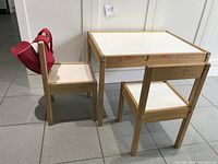 Wooden kids table with white top paired with two wooden chairs, one chair featuring an attached red heart-shaped felt pouch.