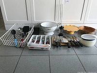 Overall view of kitchen items laid on floor featuring utensil tray with utensils, colander, cake stands, knives, and bowls