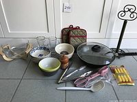 Photo showing all kitchen items including wok with lid, ceramic bowls, glass cups, metal tongs, plastic utensils, pepper grinder, pot holders, and metal paper towel holder.