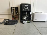 Four kitchen appliances arranged on a counter: Moulinex hand mixer in box, black Cuisinart coffee maker, white Hamilton Beach toaster, and two cast iron tortilla presses.